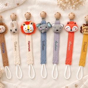 Puede incluir: Siete sujetachupetes de tela con cuentas de madera y clips metálicos. Cada uno presenta una cabeza de animal de peluche: león, conejo, león, elefante, oso, zorro y tigre. Las correas de tela están bordadas con los nombres Olivia, Victoria, Hannah, William, Andrew, Rowan y Daniel.