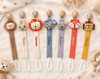 May include: Seven fabric pacifier clips with wooden beads and metal fasteners. Each features a plush animal head: lion, rabbit, lion, elephant, bear, fox, and tiger. The fabric straps are embroidered with the names Olivia, Victoria, Hannah, William, Andrew, Rowan, and Daniel.