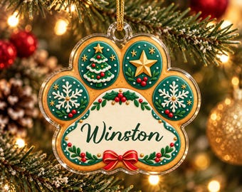 May include: A paw-shaped Christmas ornament with a clear acrylic border. The design features a tan cookie-like texture with green circular insets displaying a Christmas tree, a gold star, and white snowflakes. The center reads Winston.