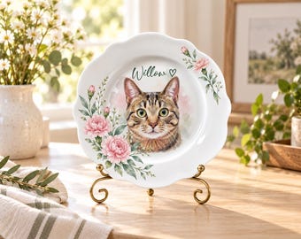 May include: A white ceramic plate with a scalloped edge featuring a watercolor illustration of a brown tabby cat, the name Willow, a small heart, and pink carnation flowers with green leaves, displayed on a gold-toned metal wire stand.