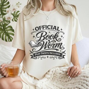 Puede incluir: Camiseta color crema con el texto "OFFICIAL Book Worm, Plot Twists Included" en escritura negra. El diseño incluye una ilustración de un libro abierto. La persona sostiene una taza de vidrio transparente con un líquido marrón.