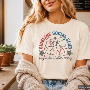 Op de afbeelding: Een crèmekleurig T-shirt met korte mouwen en een grafisch ontwerp met de tekst "SIDELINE SOCIAL CLUB" in rood en blauw, een grote strik met honkbalstiksels, geblokte sterren en de tekst "hey batter batter swing" in het zwart.