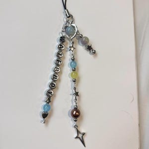 Puede incluir: Un charm de cuentas con la palabra "WATERBOY" escrita con cuentas de letras plateadas. El charm presenta varias cuentas de colores, incluyendo azul, amarillo y marrón, junto con dijes en forma de estrella y corazón. Una cuerda negra está unida en la parte superior.
