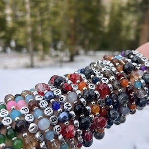 May include: A collection of beaded bracelets in various colors, including green, blue, red, and black. Some bracelets feature silver letter beads spelling out words. The bracelets are worn on a wrist, with a snowy, blurred background.
