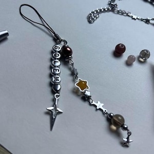 Op de afbeelding: Een zilveren telefoonbedel met het woord "GOLEM" in zilveren letterkralen. De bedel heeft een sterhanger en een ketting met ster- en edelsteenaccenten. Extra kralen en een armband zijn ook zichtbaar.