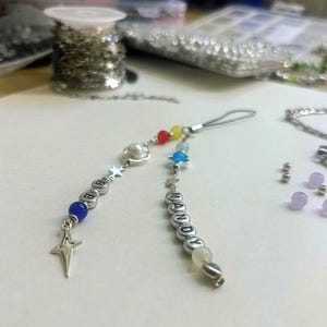 Op de afbeelding: Een handgemaakte bedel met de namen "BABY" en "MANDY" gespeld in zilveren letterkralen. De bedels hebben kleurrijke kralen, sterbedels en een parel. De achtergrond bevat spoelen met kettingen en andere knutselbenodigdheden.