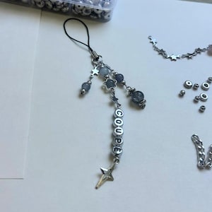 Op de afbeelding: Een handgemaakte telefoonbedel met een zwarte lus, een zilverkleurige ketting en diverse kralen. De bedel bevat grijze en blauwe kralen, stervormige bedels en letterkralen met de tekst "COUPE". Een sterbedel hangt onderaan.