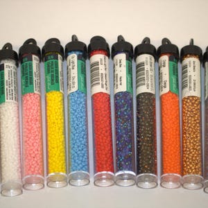 Puede incluir: Una colección de cuentas de semillas en tubos de plástico transparente. Los tubos están llenos de cuentas de varios colores, incluyendo dorado, blanco, rosa, amarillo, azul claro, rojo, morado, marrón oscuro, naranja, bronce, gris y lavanda. Cada tubo tiene una tapa negra y una etiqueta.