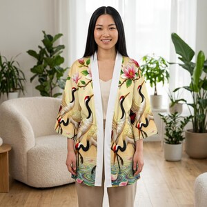 Pode incluir: Uma jaqueta estilo quimono amarelo-claro com um padrão repetitivo de grous-de-coroa-vermelha e flores de magnólia cor-de-rosa. A peça tem uma borda frontal branca sólida, mangas três quartos e um design aberto e solto.
