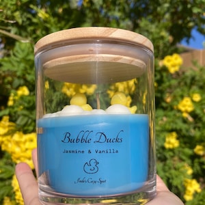 May include: A clear glass jar candle with a wooden lid. The candle is blue with the text "Bubble Ducks Jasmine & Vanilla" and a duck graphic. The candle has yellow duck and white cloud decorations.