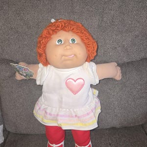May include: A vintage Cabbage Patch Kid doll with orange yarn hair, green eyes, and a white dress featuring a pink heart. The doll is wearing red leggings and white shoes. A tag is attached to the doll.