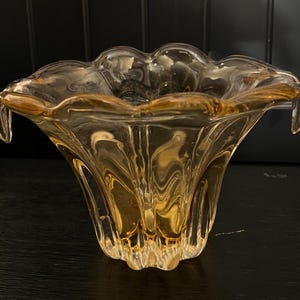 May include: A clear glass decorative bowl with a ruffled rim and a flared, petal-like shape. The bowl has a golden-amber hue and is likely made of blown glass. The bowl is sitting on a black surface.
