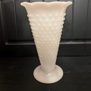 Puede incluir: Un jarrón blanco con diseño hobnail y borde festoneado. El jarrón tiene una base redonda y una superficie texturizada con pequeñas protuberancias en relieve. El jarrón es de vidrio opaco y está sobre un fondo oscuro.