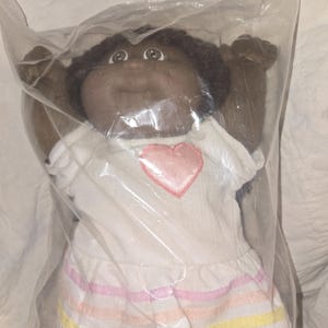 May include: A plush doll with dark skin and dark curly hair, wearing a white short-sleeved top featuring a pink heart appliqué on the chest and a skirt with horizontal pastel pink, orange, and yellow stripes, all enclosed in a clear plastic bag.