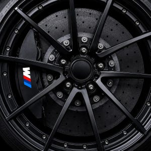 Op de afbeelding: Een matzwarte lichtmetalen velg met meerdere spaken, een centrale naafdop en een grote remklauw. De remklauw is zwart en toont het M-logo in blauw, paars en rood. Achter de spaken is een geperforeerde grijze remschijf duidelijk zichtbaar.