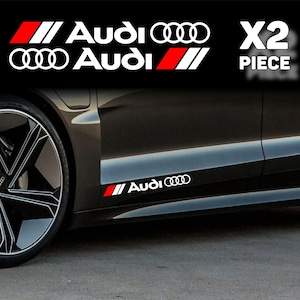 Può includere: Adesivi per auto Audi bianchi e rossi. Gli adesivi presentano il logo Audi e la scritta "Audi" in bianco, con strisce rosse e bianche. L'immagine mostra gli adesivi su un'auto nera.