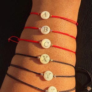 Op de afbeelding: Zes armbanden met witte, cirkelvormige bedels, elk met een andere letter van het alfabet. De armbanden zijn geregen op rode en zwarte koorden. De bedels zijn versierd met goudkleurige kralen.