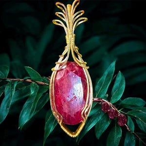 May include: A gold-toned pendant featuring a large, faceted, oval-shaped ruby. The ruby is encased in a decorative gold wire frame with an ornate top design. The pendant hangs against a backdrop of green leaves.