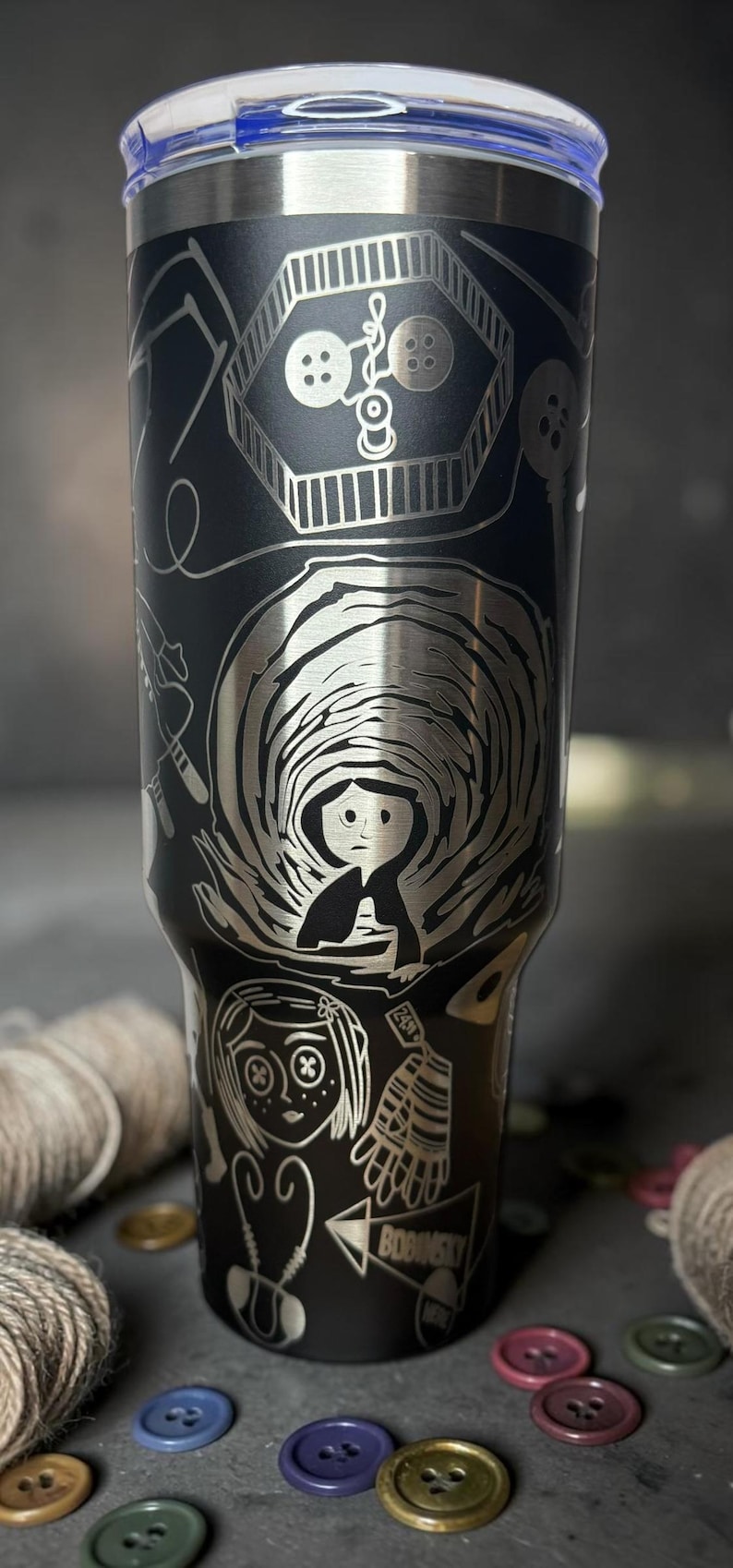 May include: Black stainless steel tumbler with a detailed engraved design. The design features imagery from a movie, including buttons, a girl, and the word "Bobinsky". The tumbler has a blue lid.