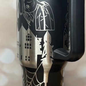 May include: Black stainless steel tumbler with a silver etched Halloween design. The design includes a witch, haunted house, spider web, and stars. The tumbler has a black handle and a blue lid.
