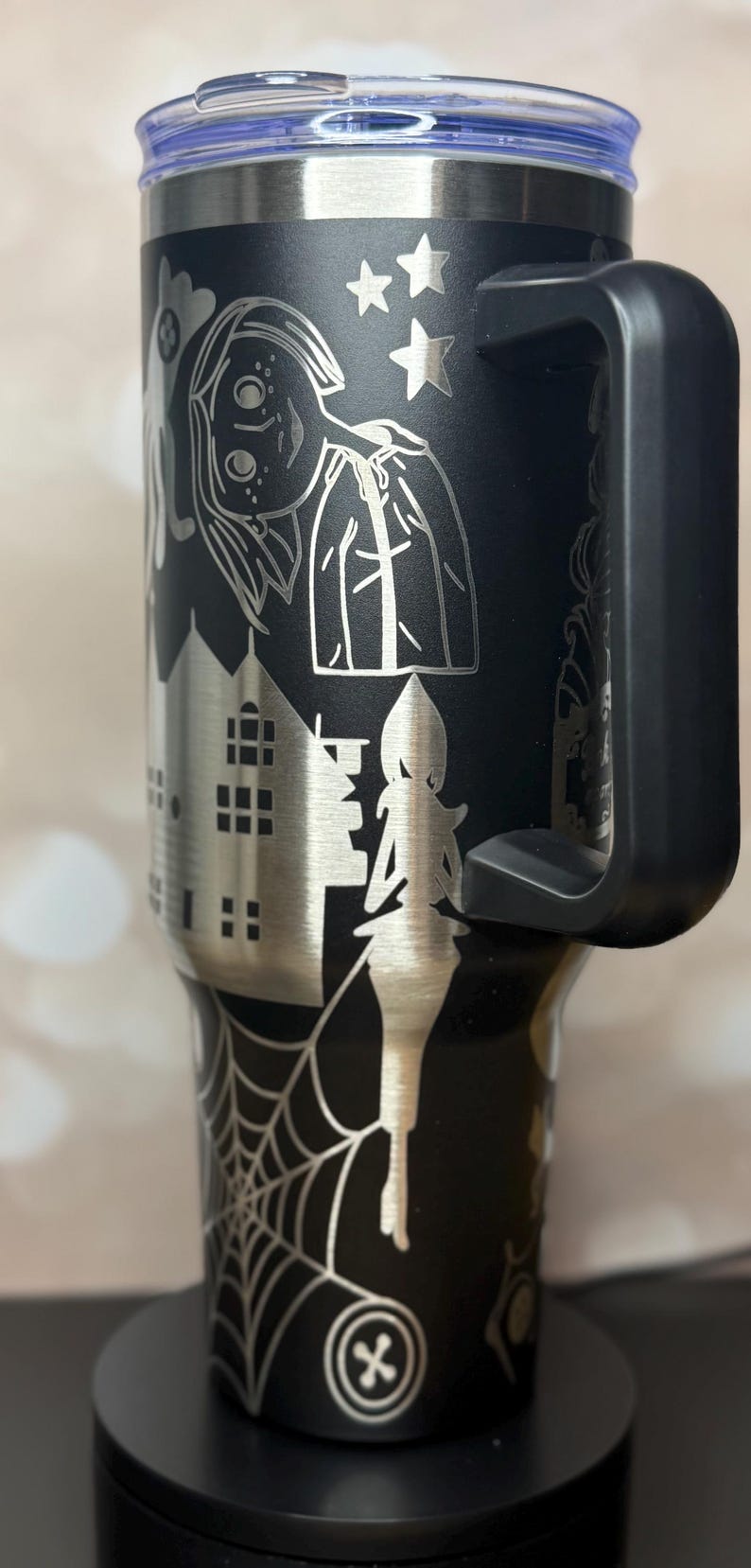 May include: Black stainless steel tumbler with a silver etched Halloween design. The design includes a witch, haunted house, spider web, and stars. The tumbler has a black handle and a blue lid.