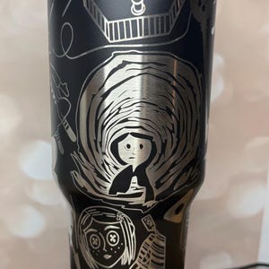 May include: Black stainless steel tumbler with a blue lid, featuring etched illustrations from a fantasy movie. The design includes a girl, buttons, and other thematic elements. A unique, themed drinkware item.