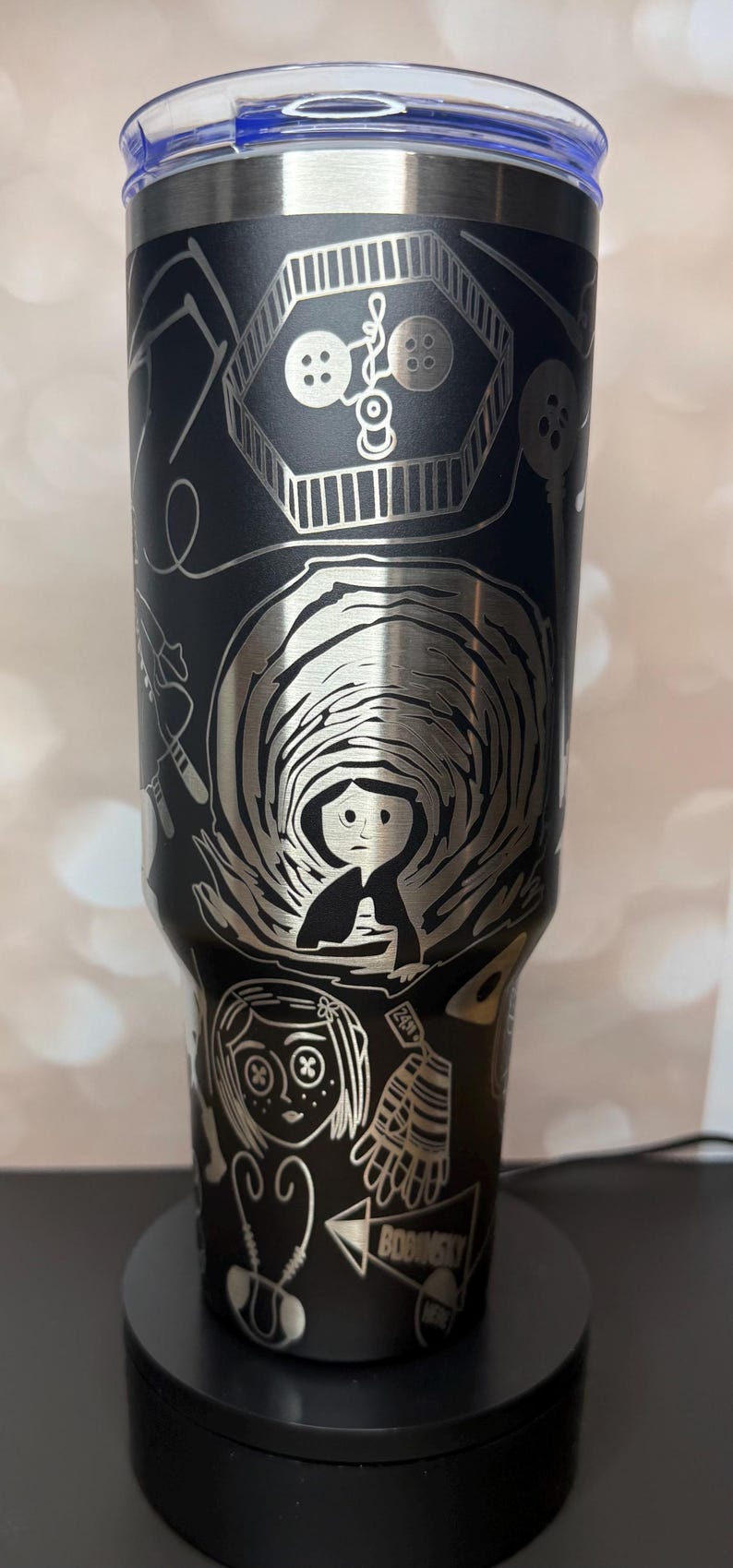 May include: Black stainless steel tumbler with a blue lid, featuring etched illustrations from a fantasy movie. The design includes a girl, buttons, and other thematic elements. A unique, themed drinkware item.