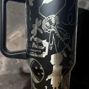 May include: Black stainless steel tumbler with a handle, featuring etched illustrations of a cartoon girl, insects, and the words "Pink Palace Apartments". The tumbler has a silver rim and a blue lid.