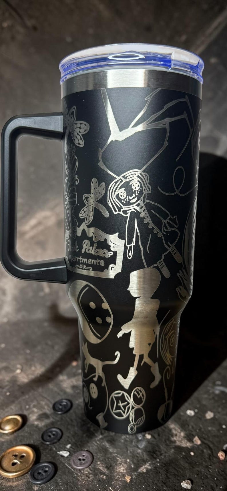 May include: Black stainless steel tumbler with a handle, featuring etched illustrations of a cartoon girl, insects, and the words "Pink Palace Apartments". The tumbler has a silver rim and a blue lid.