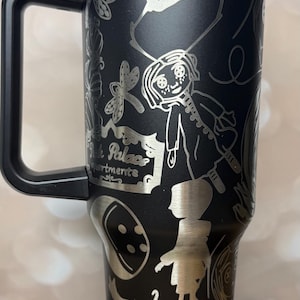 May include: Black stainless steel tumbler with a handle and a blue lid. The tumbler features etched illustrations of characters and elements from a fantasy story, including a girl, insects, and the text "Pink Palace Apartments."