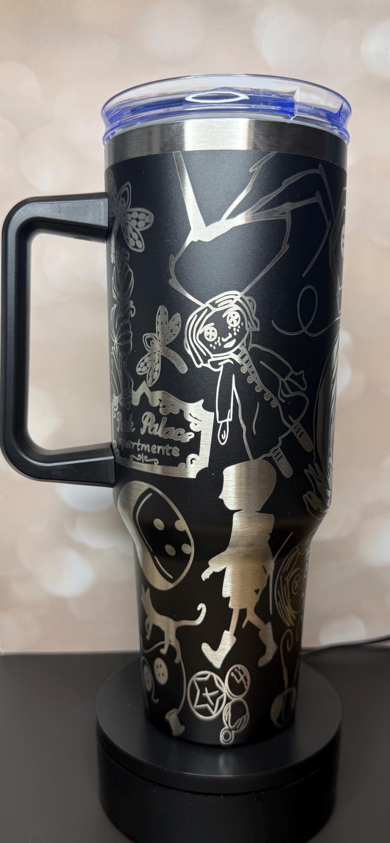 May include: Black stainless steel tumbler with a handle and a blue lid. The tumbler features etched illustrations of characters and elements from a fantasy story, including a girl, insects, and the text "Pink Palace Apartments."