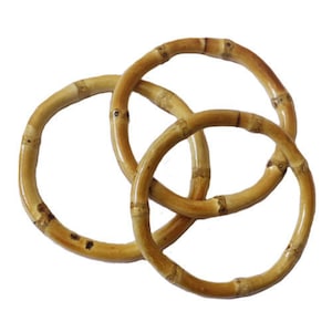 May include: Three interlocking bamboo rings, each with a natural, light brown color and a glossy finish. The rings are arranged in a decorative pattern, showcasing the natural bamboo grain and texture. The image is set against a plain white background.