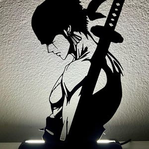 Może przedstawiać: Czarna lampa sylwetkowa przedstawiająca profil postaci z bandaną, kolczykami i mieczem katana przymocowanym do plec&oacute;w. Postać jest oświetlona od dołu przez prostokątną czarną podstawę z jasnym białym paskiem świetlnym.