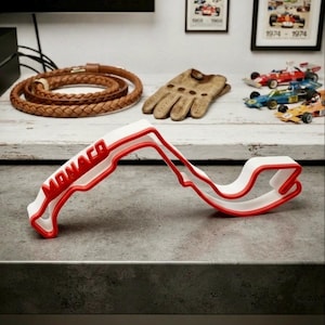 May include: A three-dimensional white and red sculpture representing the Monaco Grand Prix race track layout. The structure features the word MONACO embossed in red on the white track surface, creating a distinct outline of the circuit.