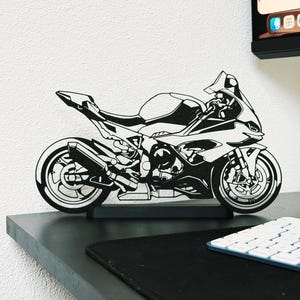 Può includere: Una sagoma ritagliata in bianco e nero di una moto sportiva montata su una base rettangolare. Il design presenta contorni dettagliati del telaio, delle ruote, del motore e delle carenature, catturando il profilo aerodinamico del veicolo.