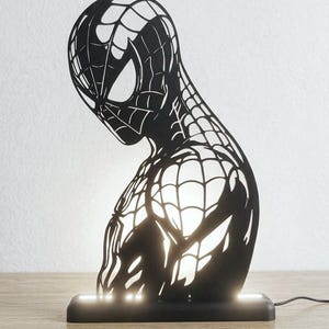 May include: A black, laser-cut silhouette lamp featuring the head and torso of the character Spider-Man. The design includes the iconic web pattern across the figure. The lamp is mounted on a rectangular black base that emits a soft white light.