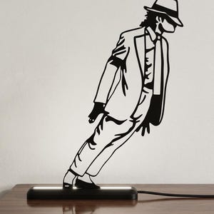 Puede incluir: Una lámpara de silueta negra que representa una figura con traje y sombrero fedora realizando un movimiento de baile inclinado hacia adelante. La figura está unida a una base rectangular negra que emite luz blanca brillante desde la parte superior.