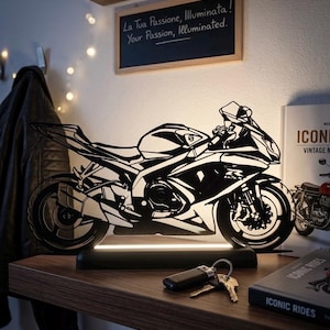 Può includere: Una lampada a LED a forma di motocicletta in bianco e nero che presenta la silhouette dettagliata di una moto sportiva. La lampada è montata su una base rettangolare nera che emette una luce soffusa, mettendo in risalto il design ritagliato.