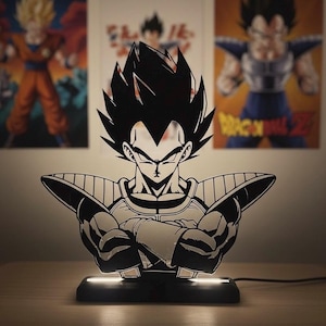 Può includere: Una lampada a LED in bianco e nero raffigurante un personaggio stilizzato con capelli a punta e braccia incrociate, montata su una base rettangolare nera che emette una luce bianca soffusa sotto la figura.