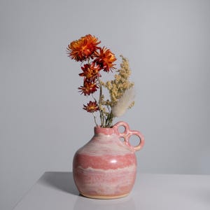 May include: A pink and white ceramic vase with two loop handles contains dried orange and yellow flowers. The vase has a rounded shape and sits on a white surface against a grey background. The flowers add a touch of colour.