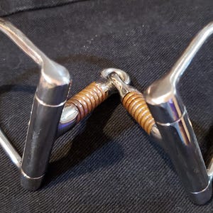 May include: A polished metal horse bit with a brown leather-wrapped center. The bit features curved side pieces and a central joint. The image is set against a dark background.