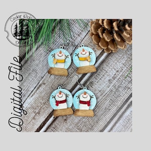Puede incluir: Un par de pendientes de madera con forma de bola de nieve con un muñeco de nieve. Las bolas de nieve están pintadas de azul con puntos blancos y presentan un muñeco de nieve con una bufanda roja o amarilla. Los pendientes están unidos a ganchos plateados.