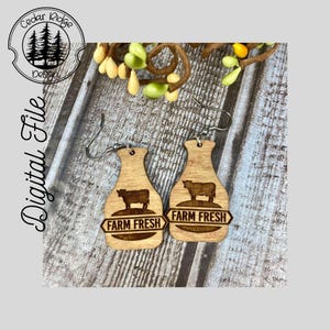 May include: A pair of wooden earrings shaped like milk bottles with a cow and the text "Farm Fresh".