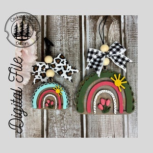 May include: Two wooden rainbow ornaments with a sun and flowers. The rainbows are layered with red, pink, and white stripes. The ornaments are decorated with black and white checkered and leopard print bows.