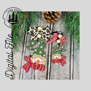 May include: Two wooden snowman ornaments with green hats, red and white scarves, and white faces. The larger ornament has a leopard print bow and the smaller ornament has a red and white gingham bow.