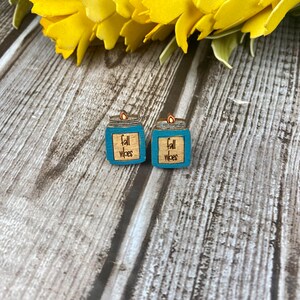 Puede incluir: Un par de pendientes de madera con forma de tarros de cristal azul verdoso con una llama en la parte superior. Los tarros tienen el texto "fall vibes" escrito en ellos.