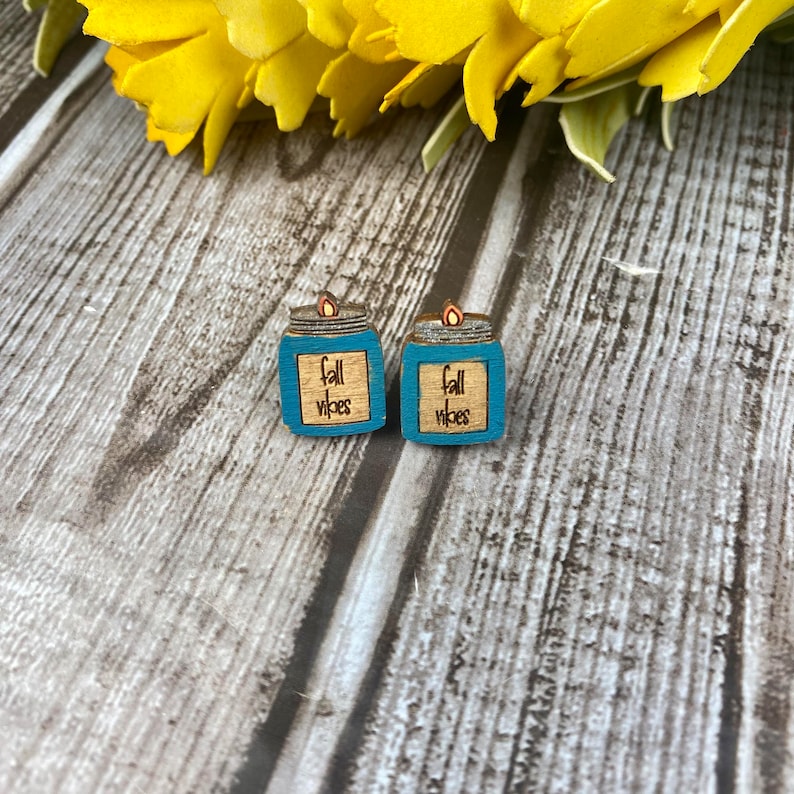 Puede incluir: Un par de pendientes de madera con forma de tarros de cristal azul verdoso con una llama en la parte superior. Los tarros tienen el texto "fall vibes" escrito en ellos.