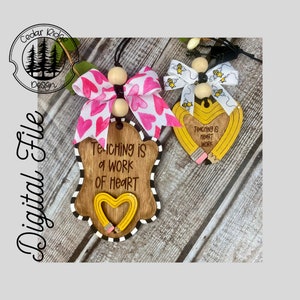 May include: Two wooden teacher appreciation ornaments. One is shield-shaped with a heart-shaped pencil and the text "Teaching is a work of heart." The other is a pencil-shaped heart with the text "Teaching is heart work." Both have bows.