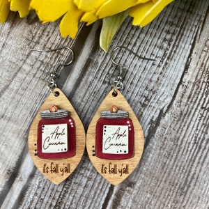 Puede incluir: Un par de pendientes de madera con un dise&ntilde;o de tarro de manzana y canela rojo y el texto "it's fall y'all".