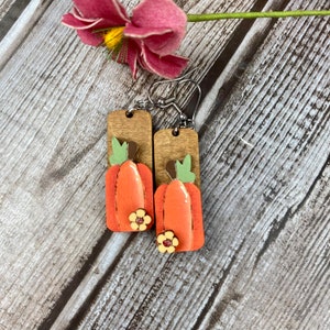 Puede incluir: Un par de pendientes de madera con un dise&ntilde;o de calabaza. Las calabazas est&aacute;n pintadas de naranja con tallos verdes y detalles marrones. Cada calabaza tiene una peque&ntilde;a flor amarilla en la parte inferior.